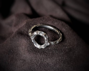 May include: A silver ring with a textured, circular design. The ring is open on one side, creating a unique and modern look.