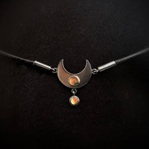 May include: A silver crescent moon pendant necklace with a small orange gemstone in the center and a dangling orange gemstone. The necklace is on a black leather cord.
