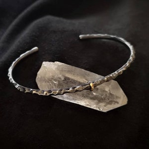 Könnte beinhalten: Ein silbernes, gehämmertes Armreif-Armband mit einem kleinen goldenen Akzent. Das Armband liegt auf einem klaren Quarzkristall.