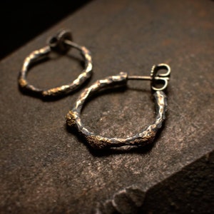 Könnte beinhalten: Ein Paar handgefertigte Silber-Creolen mit strukturierter, gehammerter Oberfläche. Die Ohrringe haben ein rustikales, organisches Design mit Akzenten aus goldfarbenem Metall. Die Ohrringe werden auf einer dunklen, strukturierten Oberfläche präsentiert.