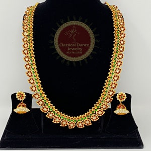 Puede incluir: Un collar de oro con piedras rojas y verdes, y pendientes a juego. El collar está en un expositor negro. Los pendientes están en un expositor negro más pequeño. El collar es de estilo tradicional indio.