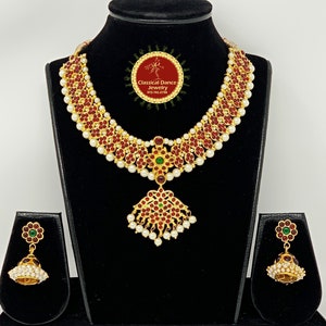 May include: A gold necklace with red and green stones and white pearls. The necklace has a pendant with a similar design. Matching earrings with red and green stones and white pearls are also included. The necklace and earrings are displayed on a black stand. The text "Classical Dance Jewelry" is visible on the stand.