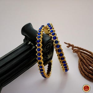May include: Two gold-colored bangles with a repeating pattern of round, deep blue gemstones. The bangles are resting on a black, textured surface. A brown tassel is visible in the background.
