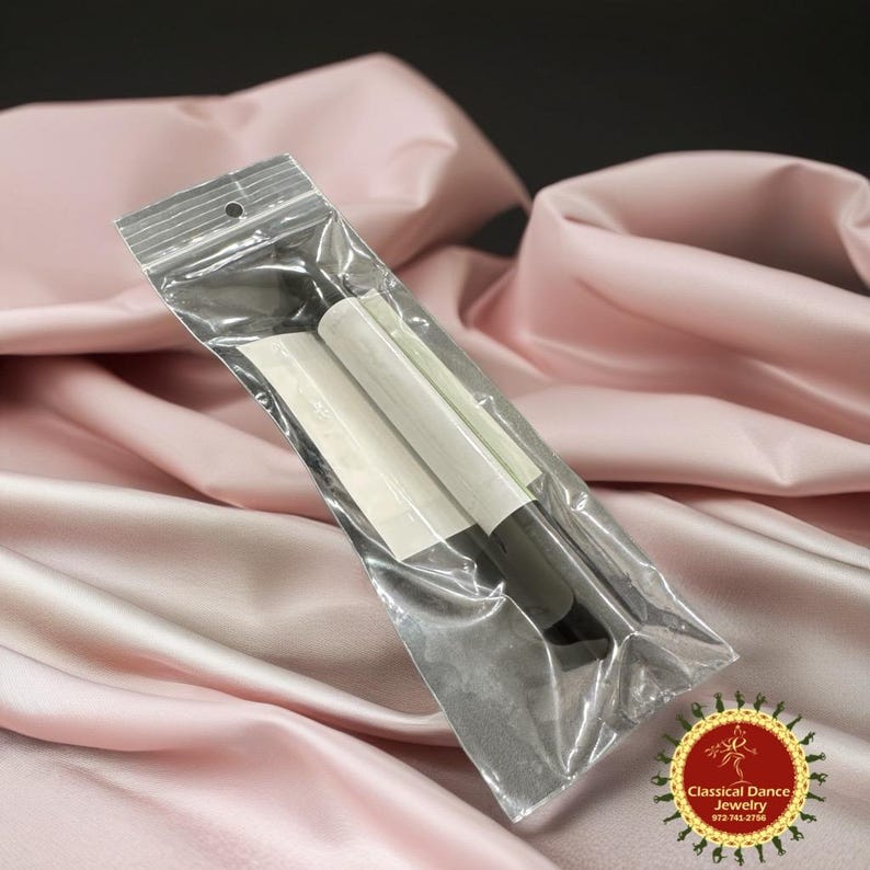 May include: A transparent plastic bag containing black dance accessories, possibly pointe shoe shanks, with white paper wrapping. The bag is sealed at the top and rests on a pink satin fabric. A gold and red logo is in the bottom right corner.