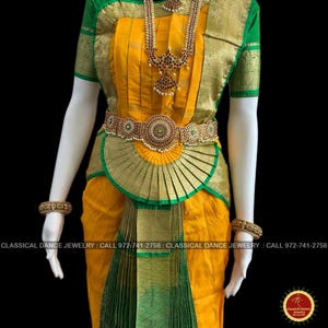May include: A yellow and green classical dance costume with gold accents. The outfit includes a pleated skirt, a decorative belt, and a long necklace with red and white stones. The sleeves are green, and the costume is displayed on a mannequin.