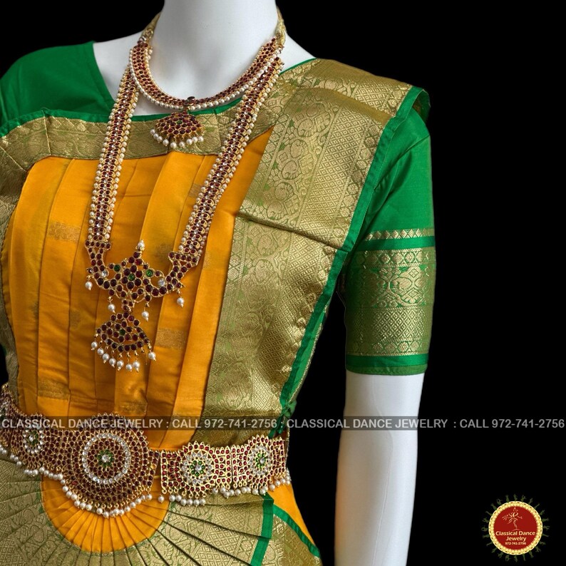 May include: A mannequin displays a traditional Indian dance costume. The ensemble features a yellow and green sari with gold accents, and ornate jewelry. The jewelry includes a necklace, long pendant, and a waist belt, all adorned with red and green stones.