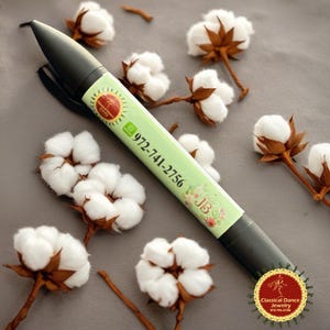 May include: A black pen with a light green label featuring the phone number 972-741-2756 and the initials JB. The pen is surrounded by cotton blossoms with white fluffy petals and brown stems, on a grey surface.