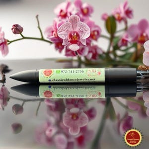May include: A black pen with a light green label featuring the text "972-741-2756" and "classicaldancejewelry.net". The pen is set against a backdrop of pink orchids, creating a visually appealing contrast.