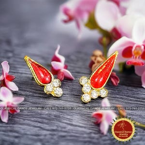 May include: A pair of gold-toned earrings with a red teardrop-shaped center and clear crystal accents. The earrings are set against a backdrop of pink flowers and a dark gray surface. The text "CLASSICAL DANCE JEWELRY" is visible.