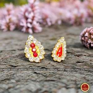 May include: A pair of gold-toned earrings with a teardrop shape. The earrings feature clear and red gemstones. The text "CLASSICAL DANCE JEWELRY" and a phone number are visible at the bottom of the image.