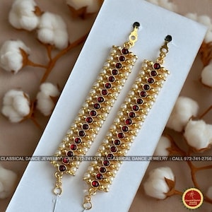 May include: Two gold-toned hair accessories with alternating rows of small white beads and red gemstones. The accessories are rectangular with a loop at the top and a small chain at the bottom. The text "CLASSICAL DANCE JEWELRY" is printed on the bottom.