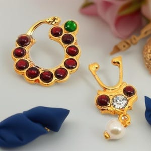 May include: Two gold-colored nose rings. One is a circular design with red and green gemstones. The other has a curved shape, red gemstones, a clear gemstone, and a dangling pearl. Jewelry.