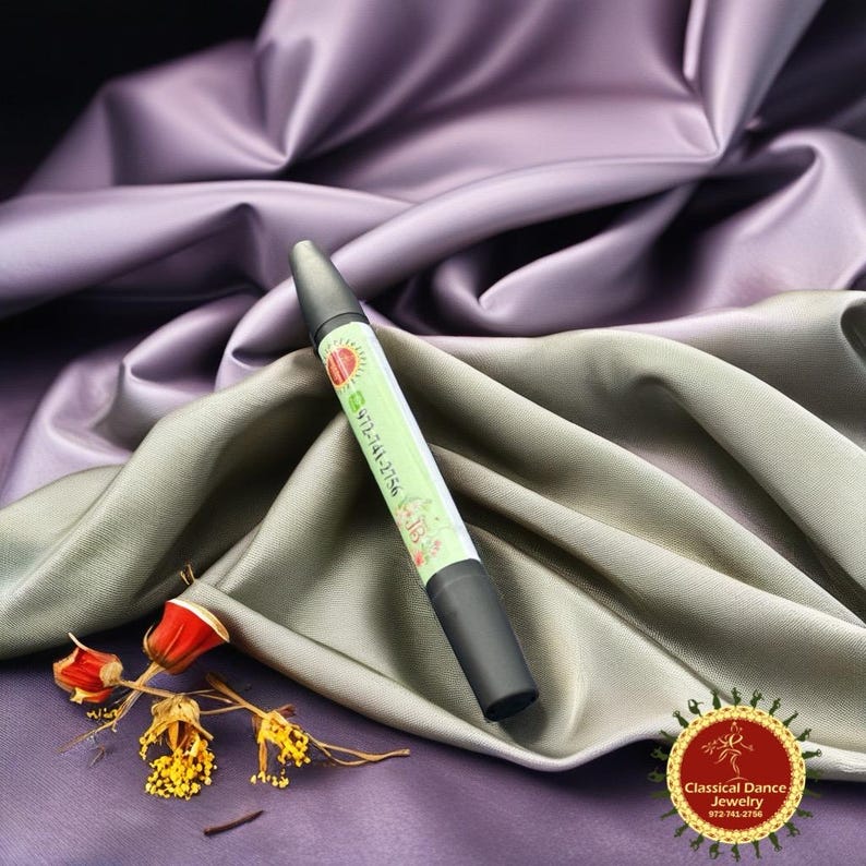 May include: A black and green eyeliner pencil with a light green label featuring a sun design and the phone number 972-741-2756. The pencil rests on draped grey and purple fabric, with dried flowers. The Classical Dance Jewelry logo is in the lower right.