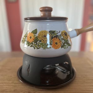 May include: A vintage enamelware pot with a wooden handle and lid. The pot is decorated with yellow and orange floral patterns on a white background. It sits on a black metal stand.