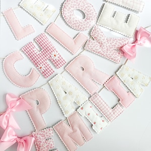 May include: A decorative banner featuring fabric letters in various shades of pink and white, including gingham, floral, and solid patterns. The letters are strung together with white cord and accented with light pink satin bows at the ends.