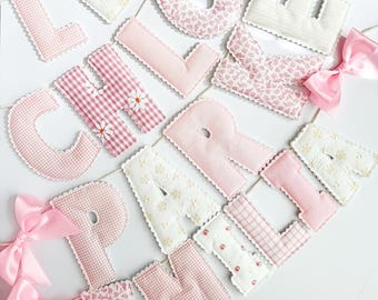 Könnte beinhalten: Ein dekoratives Banner mit Stoffbuchstaben in verschiedenen Rosa- und Weißtönen, darunter Vichy-Karo-, Blumen- und einfarbige Muster. Die Buchstaben sind mit einer weißen Kordel verbunden und an den Enden mit hellrosa Satinschleifen verziert.