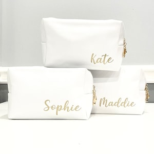 K&ouml;nnte beinhalten: Drei wei&szlig;e Rei&szlig;verschlusstaschen mit goldenen Rei&szlig;verschl&uuml;ssen und goldenen Beschriftungen. Die Taschen sind &uuml;bereinander gestapelt. Die oberste Tasche tr&auml;gt die Aufschrift "Kate", die mittlere Tasche die Aufschrift "Sophie" und die unterste Tasche die Aufschrift "Maddie".