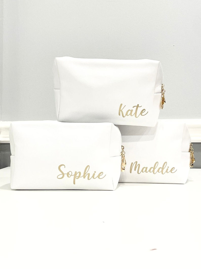 K&ouml;nnte beinhalten: Drei wei&szlig;e Rei&szlig;verschlusstaschen mit goldenen Rei&szlig;verschl&uuml;ssen und goldenen Beschriftungen. Die Taschen sind &uuml;bereinander gestapelt. Die oberste Tasche tr&auml;gt die Aufschrift "Kate", die mittlere Tasche die Aufschrift "Sophie" und die unterste Tasche die Aufschrift "Maddie".