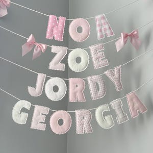 May include: A decorative banner featuring four rows of soft, fabric-covered letters spelling out the names NOA, ZOE, JORDY, and GEORGIA. The letters are in shades of pink and white with various patterns, accented by small pink satin bows on white string.