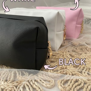 K&ouml;nnte beinhalten: Drei Kosmetiktaschen in Schwarz, Wei&szlig; und Rosa. Die Taschen sind aus einem strukturierten Material gefertigt und haben goldene Rei&szlig;verschl&uuml;sse. Die Taschen liegen auf einer gewebten Oberfl&auml;che.