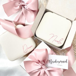 May include: Three white gift boxes with pink satin ribbon bows. The boxes are personalised with the names "Dawn", "Nicola", and "Maid of Honour Delaney". One box also has a tag that says "Bridesmaid Leagh".