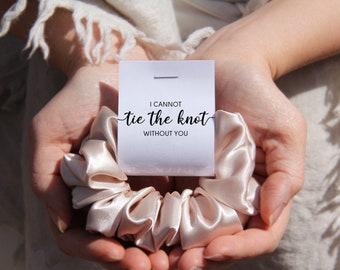 May include: A satin scrunchie in a light champagne color, with a small white card that reads "I cannot tie the knot without you". The scrunchie is held in open hands, suggesting a wedding or bridal theme.