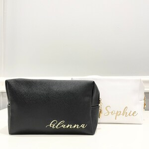 K&ouml;nnte beinhalten: Zwei schwarze und wei&szlig;e Kosmetiktaschen mit Rei&szlig;verschluss und goldener Beschriftung. Die schwarze Tasche tr&auml;gt den Schriftzug "Alanna" und die wei&szlig;e Tasche den Schriftzug "Sophie".