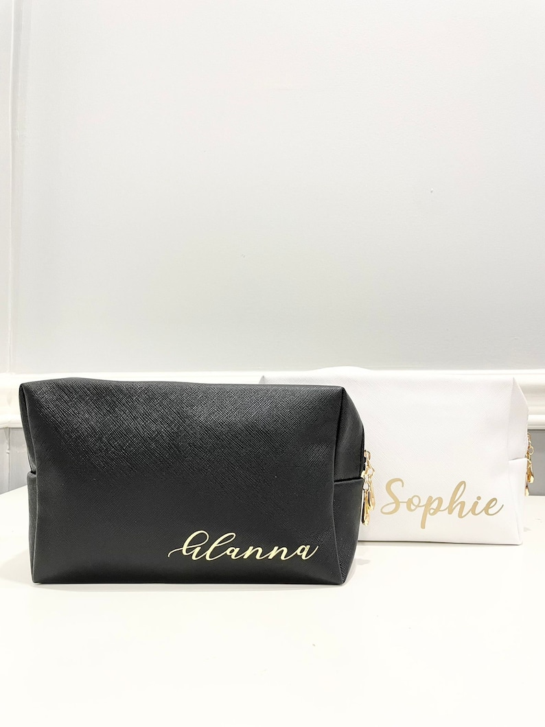 K&ouml;nnte beinhalten: Zwei schwarze und wei&szlig;e Kosmetiktaschen mit Rei&szlig;verschluss und goldener Beschriftung. Die schwarze Tasche tr&auml;gt den Schriftzug "Alanna" und die wei&szlig;e Tasche den Schriftzug "Sophie".
