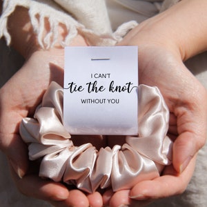 May include: A satin scrunchie in a light champagne color is held in open hands. A small white card is attached to the scrunchie with the text "I can't tie the knot without you" in black script. The background is a soft, neutral color.