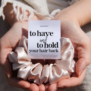 May include: A light beige satin scrunchie with the text "to have and to hold your hair back" on a white card.