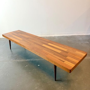 May include: A long, rectangular wooden coffee table with a warm brown finish. The tabletop is constructed from rectangular wood pieces arranged in a pattern. The table has four dark, tapered legs. The table is approximately 60 inches long.