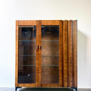 May include: A rectangular wooden display cabinet featuring two glass doors with small vertical handles. The cabinet has a rich brown wood grain finish and a textured, patterned interior back panel. It stands on four short, dark-colored metal legs.
