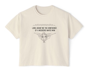 May include: A cream colored t-shirt with a graphic print of an angel with the text "LORD, GRANT ME THE CONFIDENCE OF A MEDIOCRE WHITE MAN".