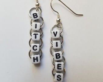 May include: A pair of silver-tone earrings. Each earring features a series of small, white square beads spelling out "BITCH VIBES" in black letters. The beads are connected by silver rings and hang from a silver fish hook.