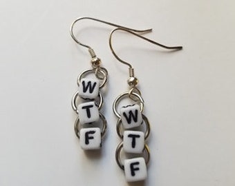May include: A pair of silver-tone earrings. Each earring features three white square beads with black letters spelling "WTF". The beads are connected by small silver rings and hang from a silver fish hook.