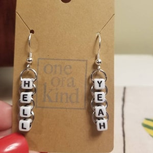 Può includere: Orecchini color argento con le parole "HELL YEAH" scritte con perline bianche. Gli orecchini sono esposti su una carta marrone con il testo "one of a Kind".