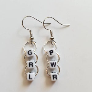 May include: Silver-tone dangle earrings. Each earring features three white square beads with black letters spelling "GRL PWR". The beads are connected by silver rings and attached to a silver fish hook ear wire. The earrings are displayed on a white surface.