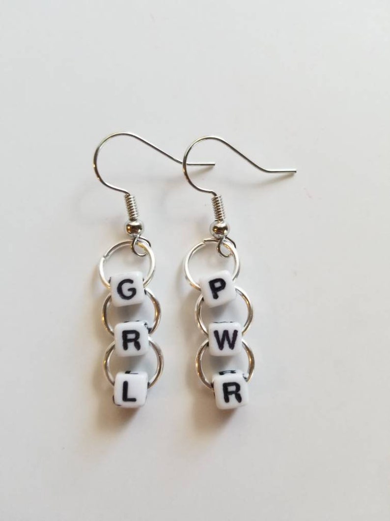 May include: Silver-tone dangle earrings. Each earring features three white square beads with black letters spelling "GRL PWR". The beads are connected by silver rings and attached to a silver fish hook ear wire. The earrings are displayed on a white surface.