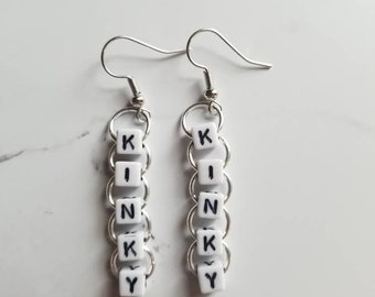 May include: A pair of silver-tone earrings. Each earring features a series of white square beads with black letters spelling out the word "KIN-KY". The beads are connected by silver rings and hang from a silver fish hook.