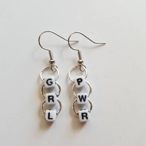 May include: A pair of silver-toned earrings. Each earring features three white square beads with black letters spelling "GRL" and "PWR". The beads are connected by silver rings and hang from a silver hook.