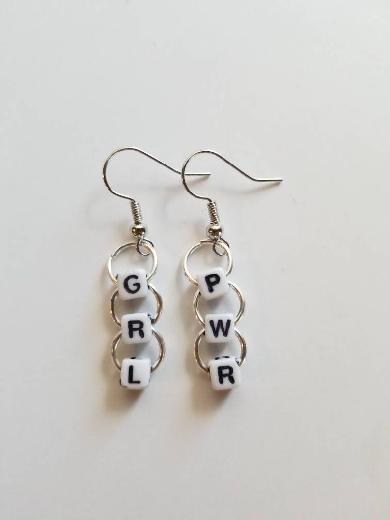 May include: A pair of silver-toned earrings. Each earring features three white square beads with black letters spelling "GRL" and "PWR". The beads are connected by silver rings and hang from a silver hook.