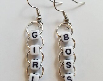 May include: A pair of dangle earrings with the words "GIRL BOSS" spelled out in black letters on white square beads. The beads are connected by silver rings and hang from silver fish hook ear wires.