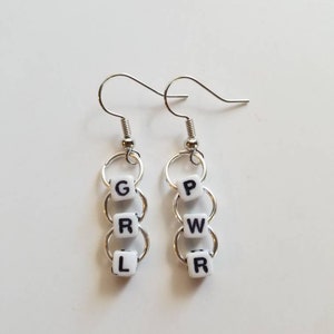 May include: Silver-tone dangle earrings. Each earring features three white square beads with black letters spelling "GRL PWR". The beads are connected by silver rings and hang from a silver fish hook.