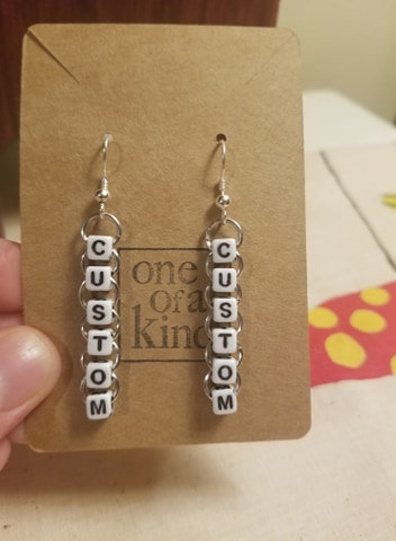 Peut inclure: Une paire de boucles d'oreilles personnalis&eacute;es avec le mot "CUSTOM" &eacute;pel&eacute; en perles de lettres blanches. Les boucles d'oreilles sont suspendues &agrave; des crochets argent&eacute;s et reli&eacute;es par de petits anneaux argent&eacute;s. Elles sont pr&eacute;sent&eacute;es sur une carte marron.