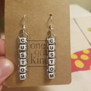 Peut inclure: Boucles d'oreilles personnalis&eacute;es avec des perles blanches &eacute;pelant "CUSTOM", suspendues &agrave; des crochets argent&eacute;s. Elles sont pr&eacute;sent&eacute;es sur une carte marron avec le texte "one of a kind".