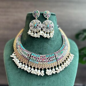 Puede incluir: Un collar de plata y oro con un diseño de piedras de colores y detalles de perlas. El collar se combina con pendientes a juego.