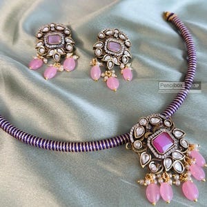 May include: A purple and gold necklace with a pink stone pendant and matching earrings. The necklace has a purple cord with gold accents and the pendant is surrounded by smaller stones. The earrings have a similar design with a pink stone and smaller stones.