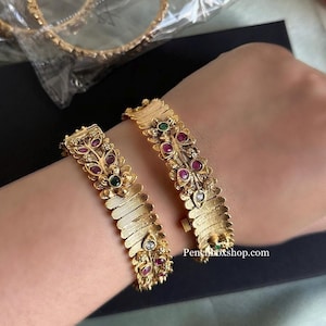 May include: Two gold bangles with intricate floral designs and red, green, and white gemstones. The bangles are worn on a person's wrist.
