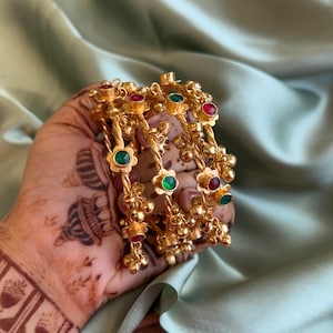 May include: A set of gold-toned bangles featuring a twisted metal band design. Each bangle is adorned with small, alternating flower-shaped settings containing round red and green stones, interspersed with tiny dangling gold-toned metal beads.