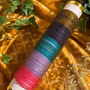 Könnte beinhalten: Eine Sammlung farbenfroher Armreifen in Pink, Lila, Türkis, Blau und Gold. Die Armreifen sind auf einem weißen Zylinder mit goldenen Akzenten gestapelt, vor einem strukturierten gelben Hintergrund mit Grün.
